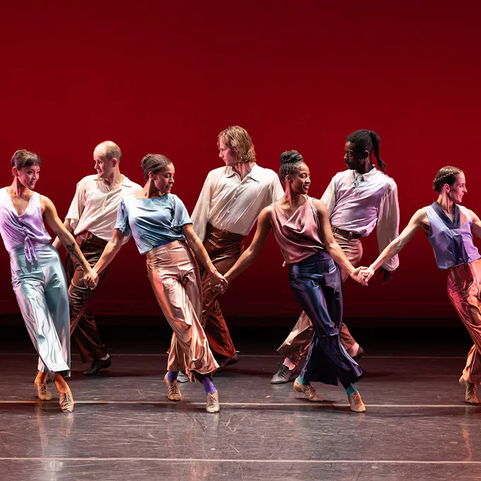 Four female dancers hold hands in a line on stage as four male dancers strike the opposite pose behind them