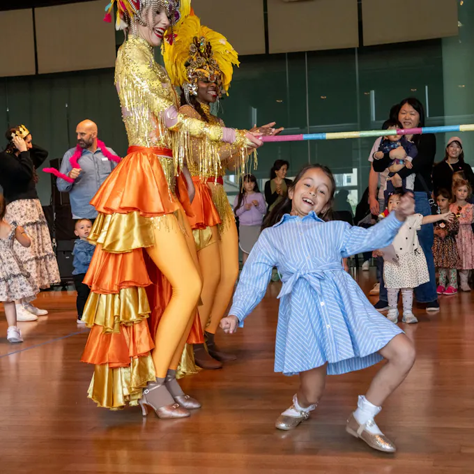 A little girl in a blue striped dress limbos under a pole held by a woman in a colorful orange and yellow costume