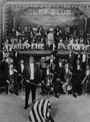 Members of the Clef Club Orchestra seated with instruments and James Reese Europe standing in front, 1912