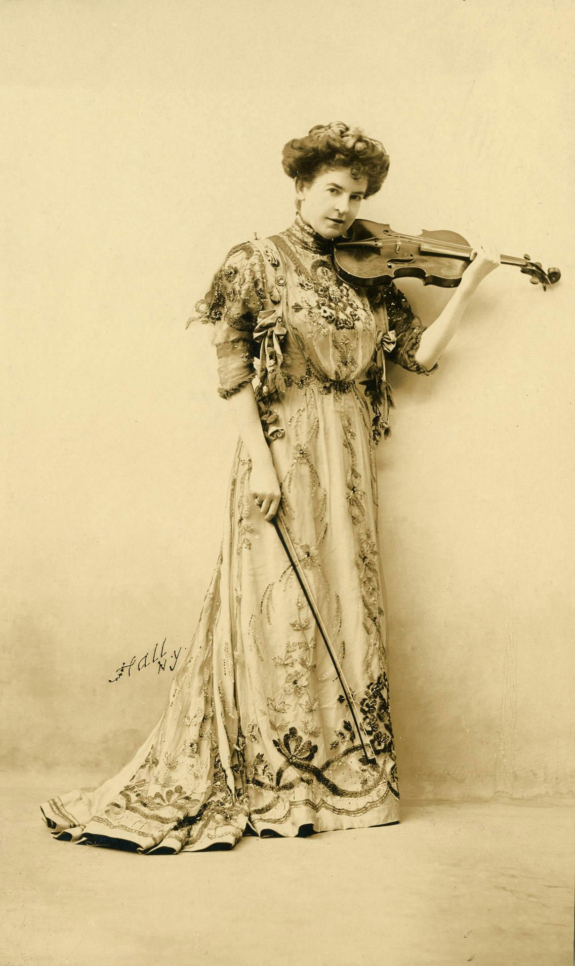 Sepia full-body photo of Maud Powell in an elaborate embroidered dress holding her violin
