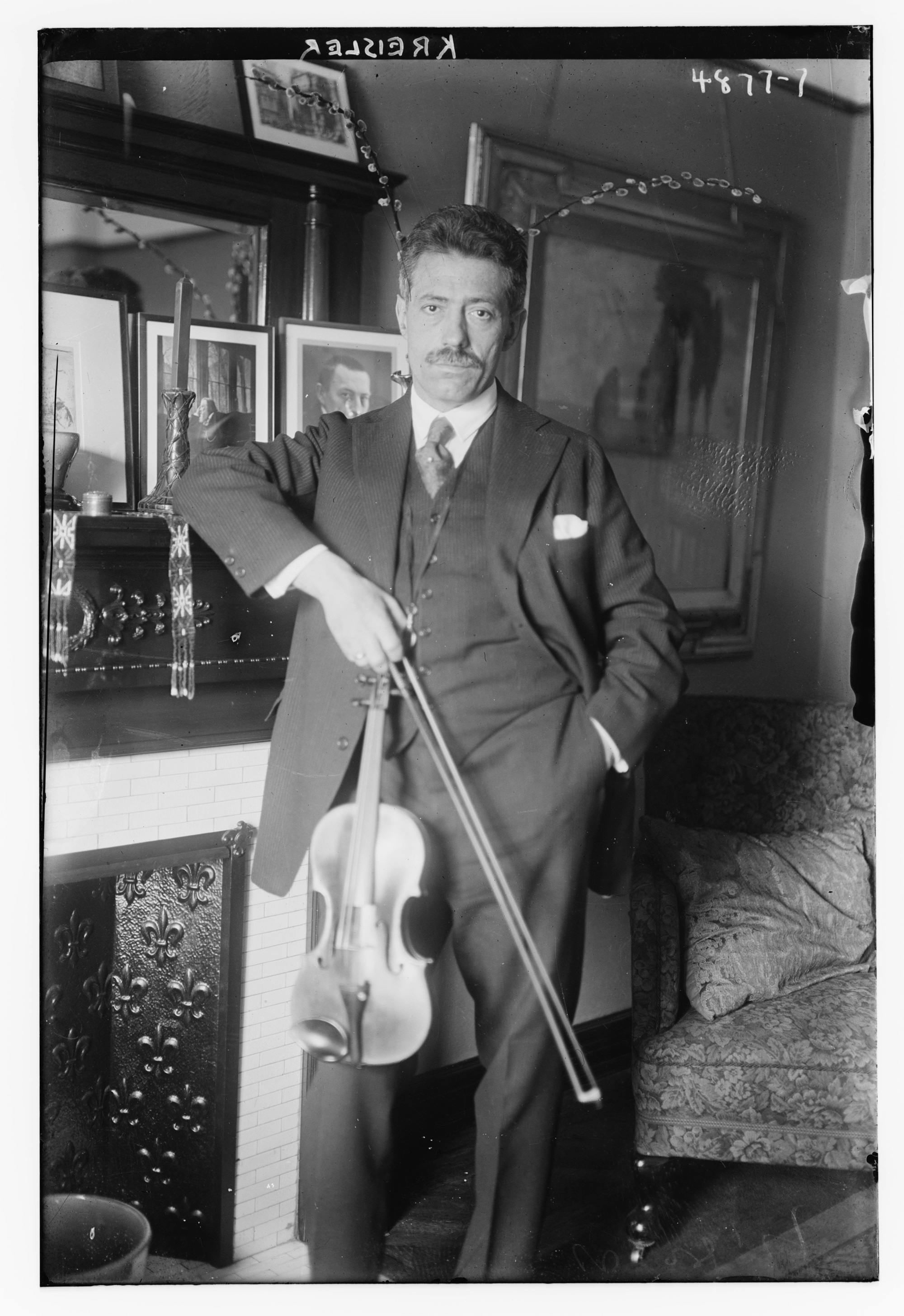 Black-and-white photo of Fritz Kreisler leaning on a mantel in a living room, holding his violin