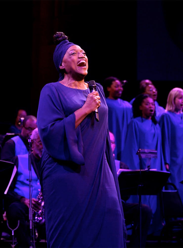 Jessye Norman is wearing a blue blouse and headband, standing in front of a chorus singing into a microphone.