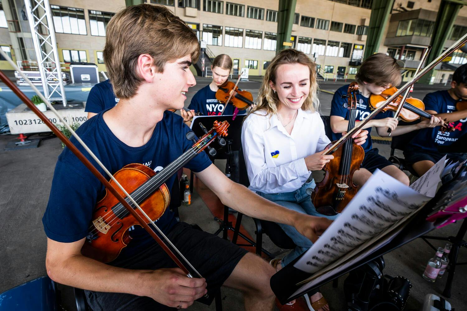 A young male and female violinist smile over their sheet music in a rehearsal room