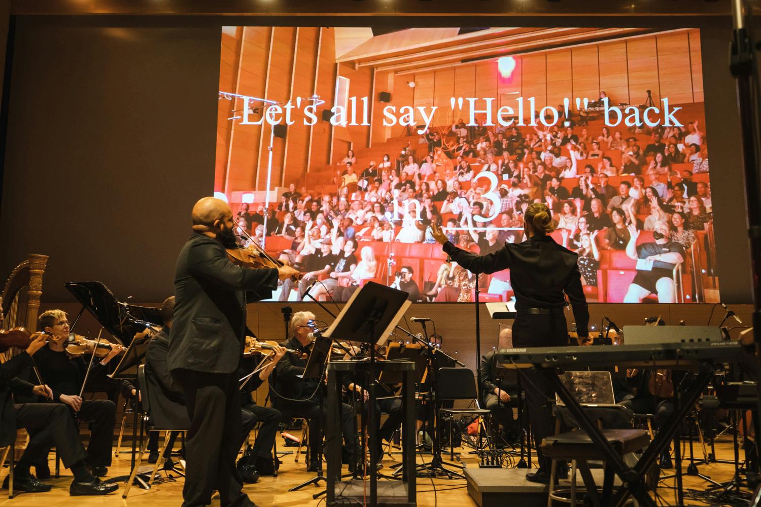 Trever New playing the violin with the orchestra in front of a projection screen that includes the text “Let’s all say ‘Hello!’ back”