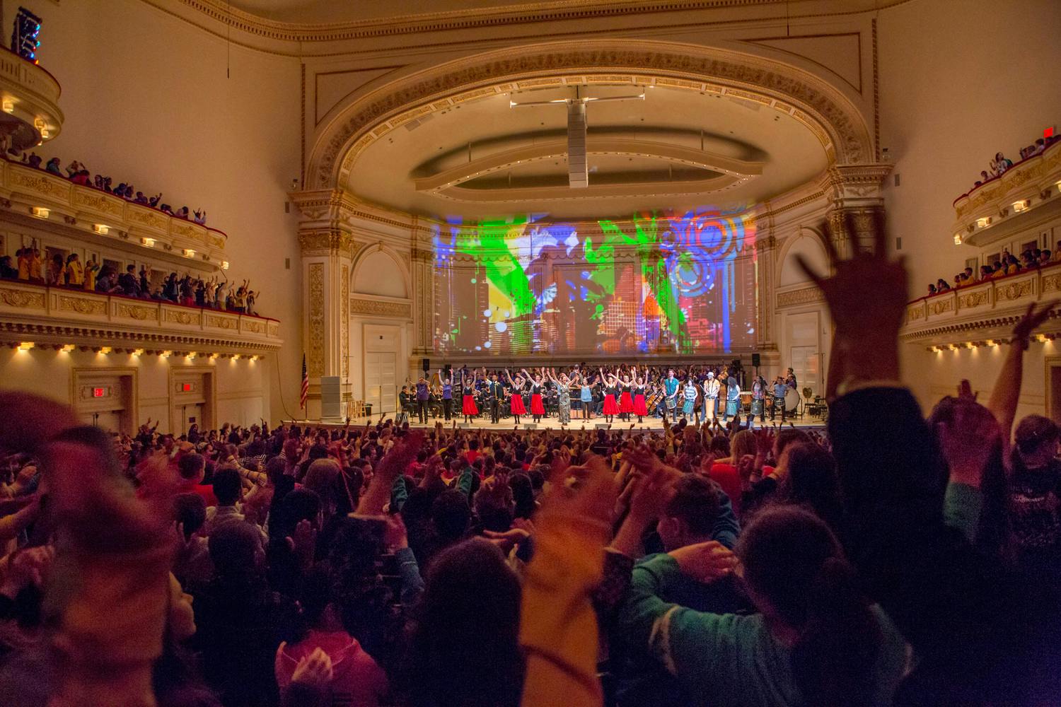 Stern Auditorium / Perelman Stage filled with audiences raising their hands at Carnegie Hall, with a colorful projection of art and dancers on the stage