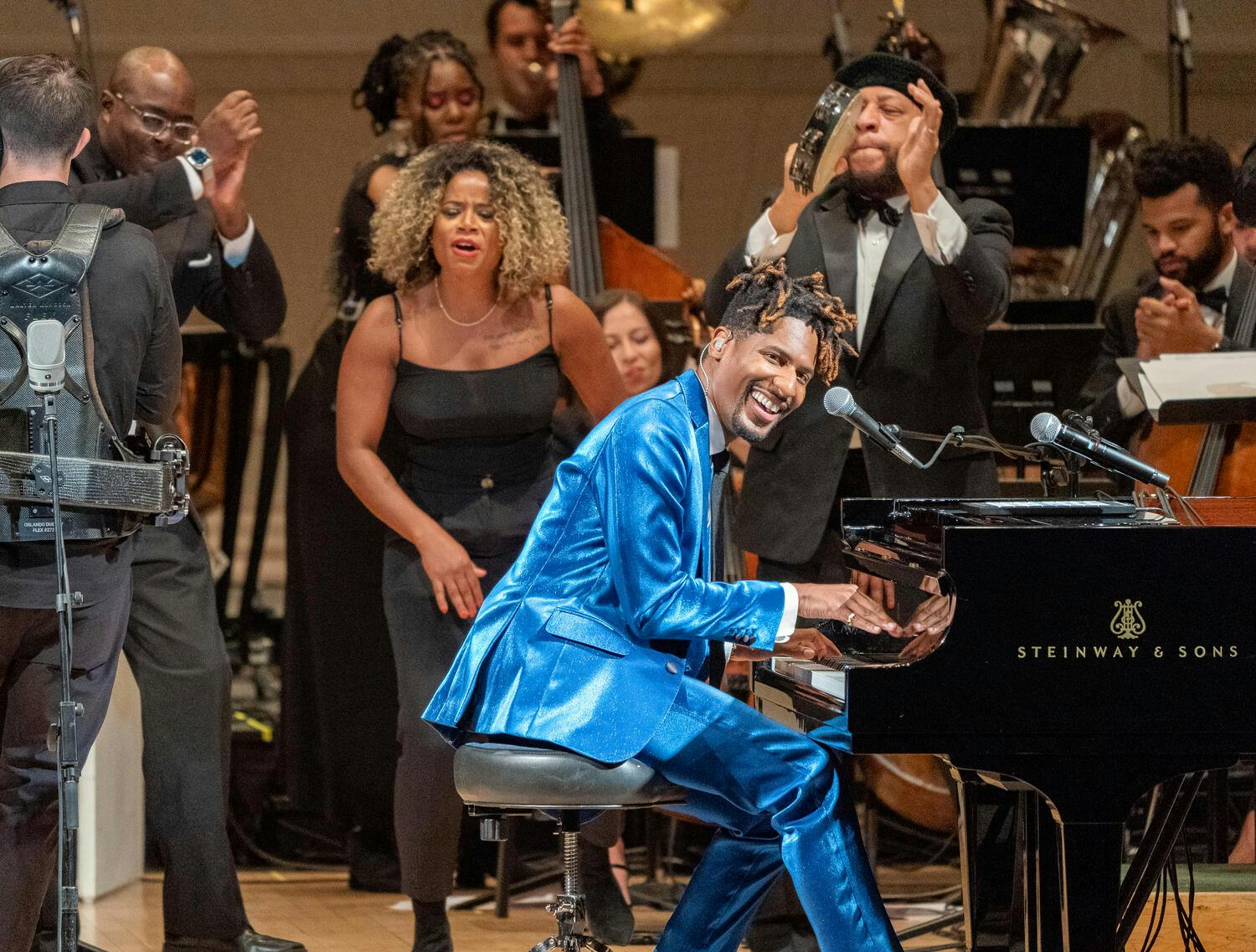 Jon Batiste playing the grand piano with backup singers and musicians on stage.
