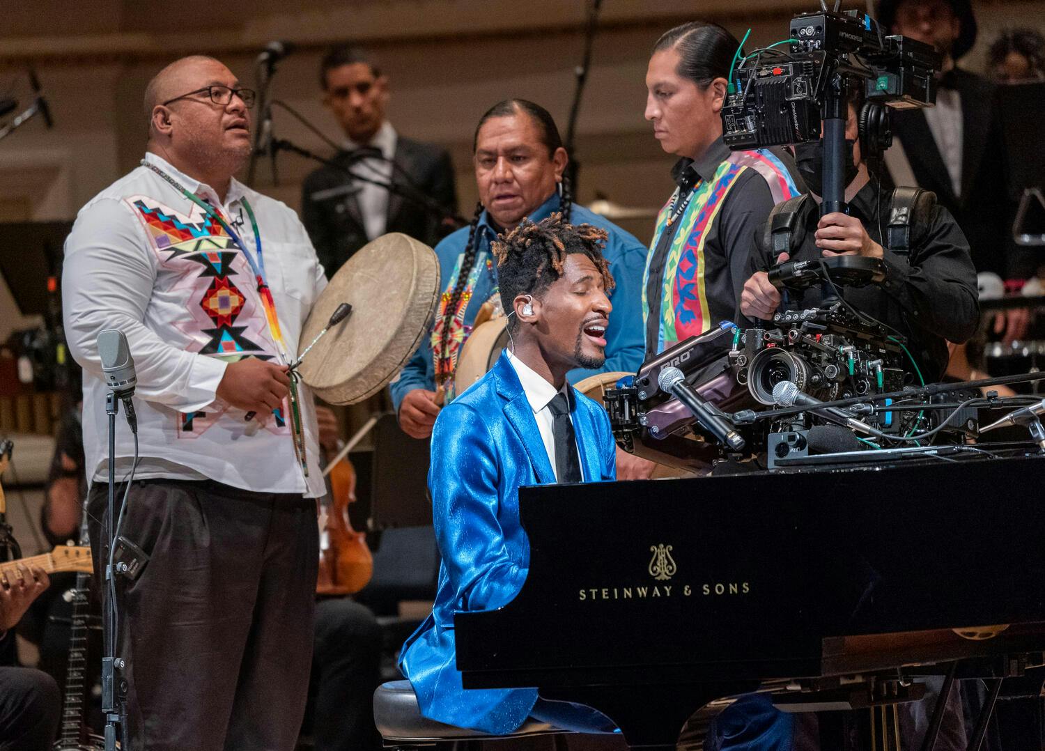 Jon Batiste playing and singing on the grand piano with a group of musicians in the back.