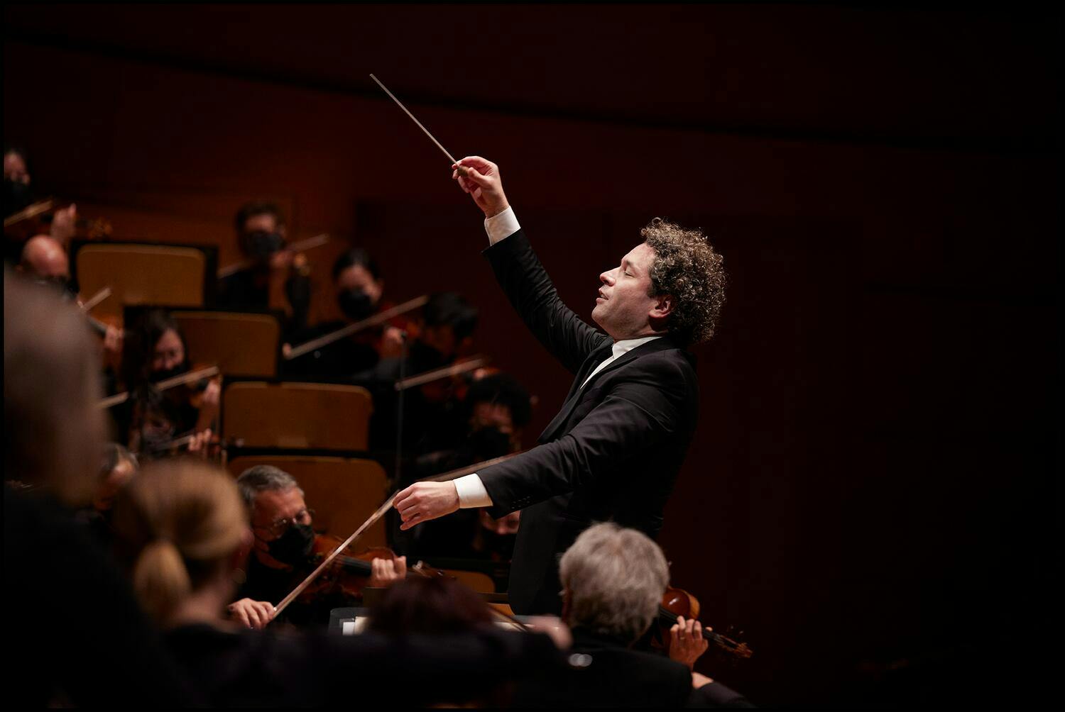 Gustavo Dudamel conducting an orchestra while holding a baton with his right arm raised.