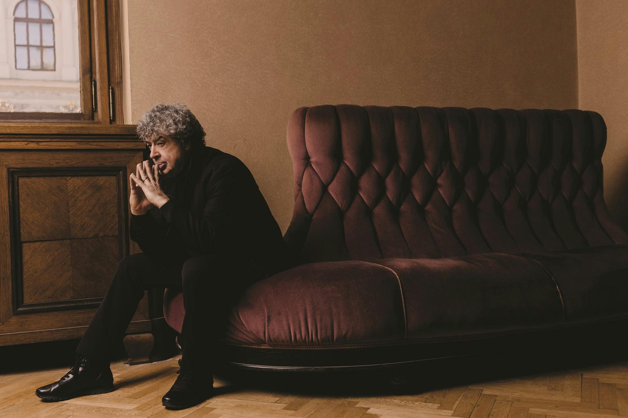 Semyon Bychkov sitting sideways on a sofa with his hands clasped by his face.