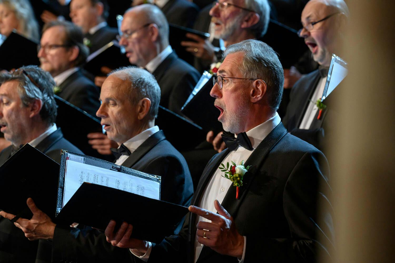 Members of the Prague Philharmonic Choir singing with their music folders open at a concert