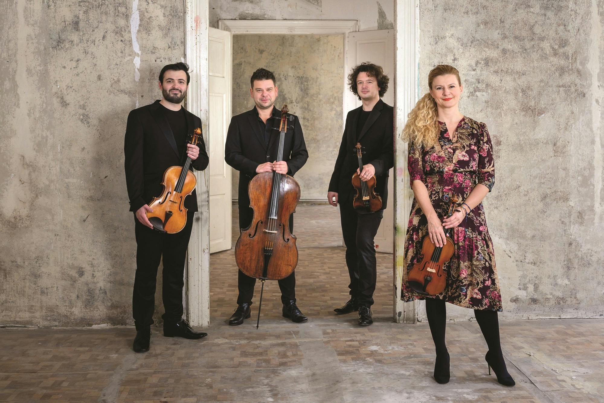 Members of the Pavel Haas Quartet standing with their instruments by a concrete wall.