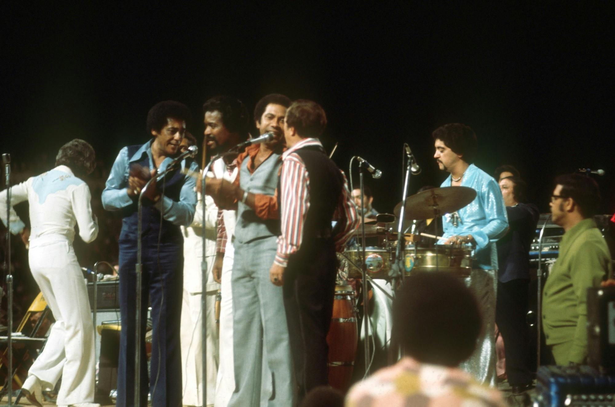 Members of the Fania All-Stars singing on stage with instruments.