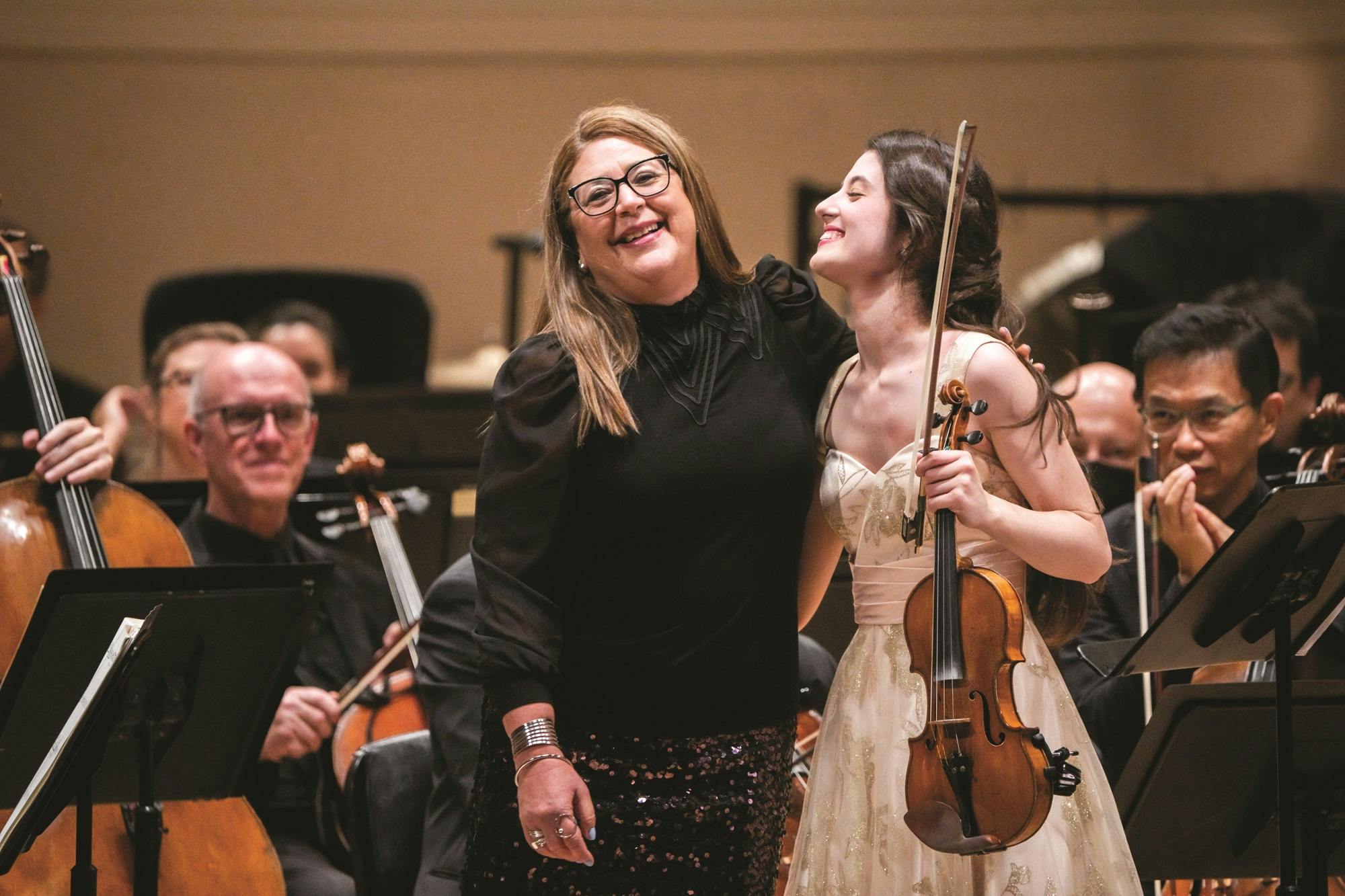 Gabriela Ortiz and Maria Dueñas standing side by side with the members of the Los Angeles Philharmonic sitting on stage behind them. 