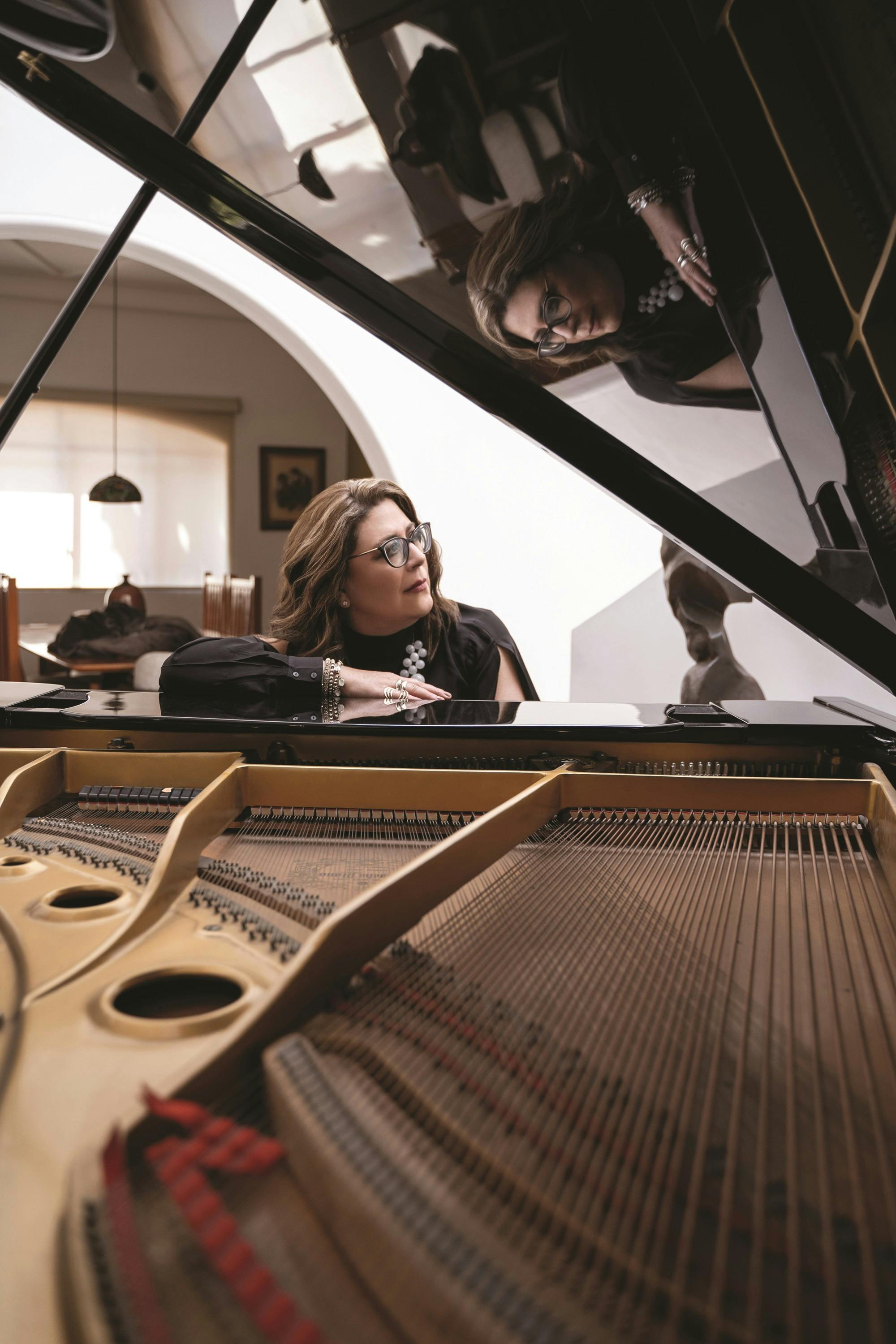 Gabriela Ortiz looking to the side while sitting at an open piano with her reflection showing on the piano lid.