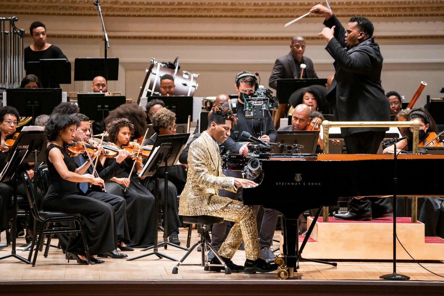 Jon Batiste playing piano on stage at Carnegie Hall with conductor Anthony Parnther and the Gateways Festival Orchestra