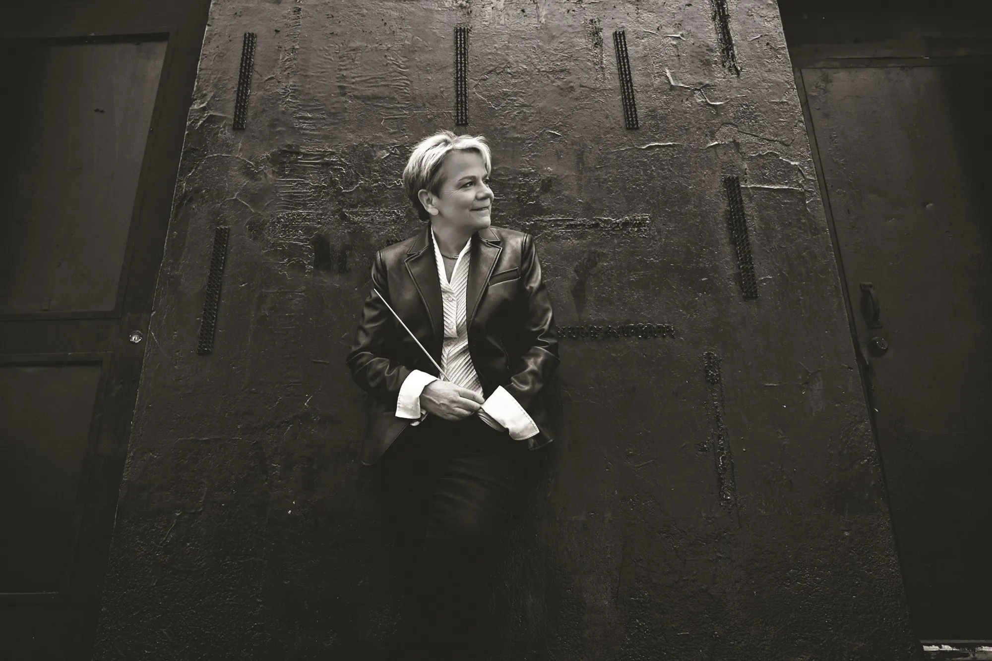 Marin Alsop holding a baton and leaning against a wall.