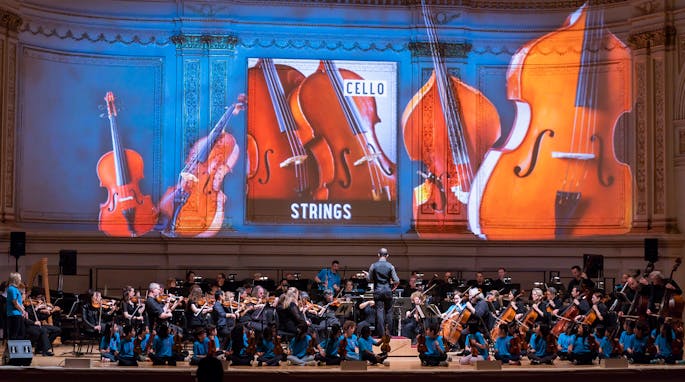 Images of cellos projected on the wall behind an orchestra
