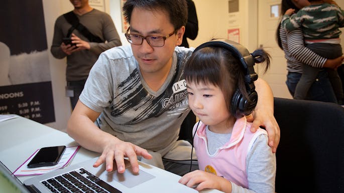A man with glasses works on a laptop with a young girl wearing headphones and a pink shirt.