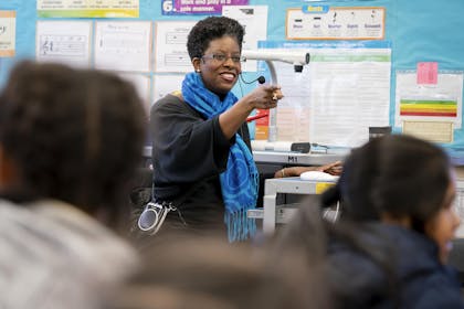 In an elementary classroom, a woman with a thick blue scarf smiles and points toward children. 