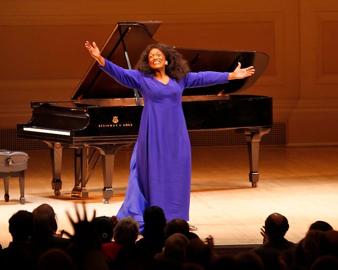 Jessye Norman wearing a purple gown stands onstage with arms outstretched as an audience applauds