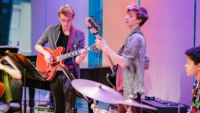 Three Future Music Ensemble members perform on stage with electric guitars and a drum set.