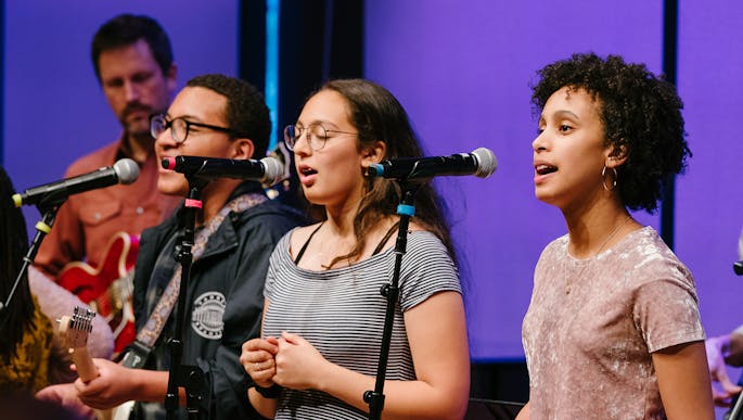 Future Music Project Ensemble members sing together in front of mic stands and purple lighting.