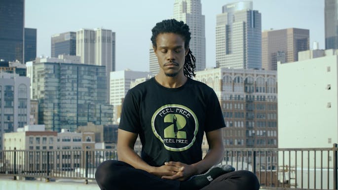 A young man sits with eyes closed in a meditative pose, a cityscape behind him