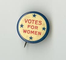 A pin with a blue border and yellow background, with four blue stars and text in red font that says, “Votes for Women”
