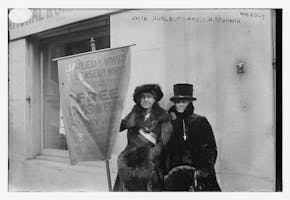 Black-and-white photograph of suffragists Julia S. Hurlbut and Eunice Dana Brannan standing in front of the National Woman’s Party headquarters in New York City, holding banners announcing a mass meeting on January 4, 1918, at Carnegie Hall.