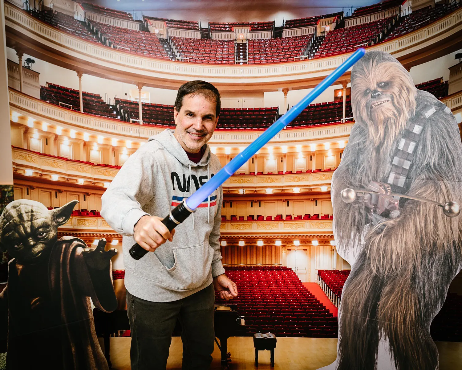 A man in a grey sweatshirt holds a blue lightsaber with Star Wars character cutouts behind him.