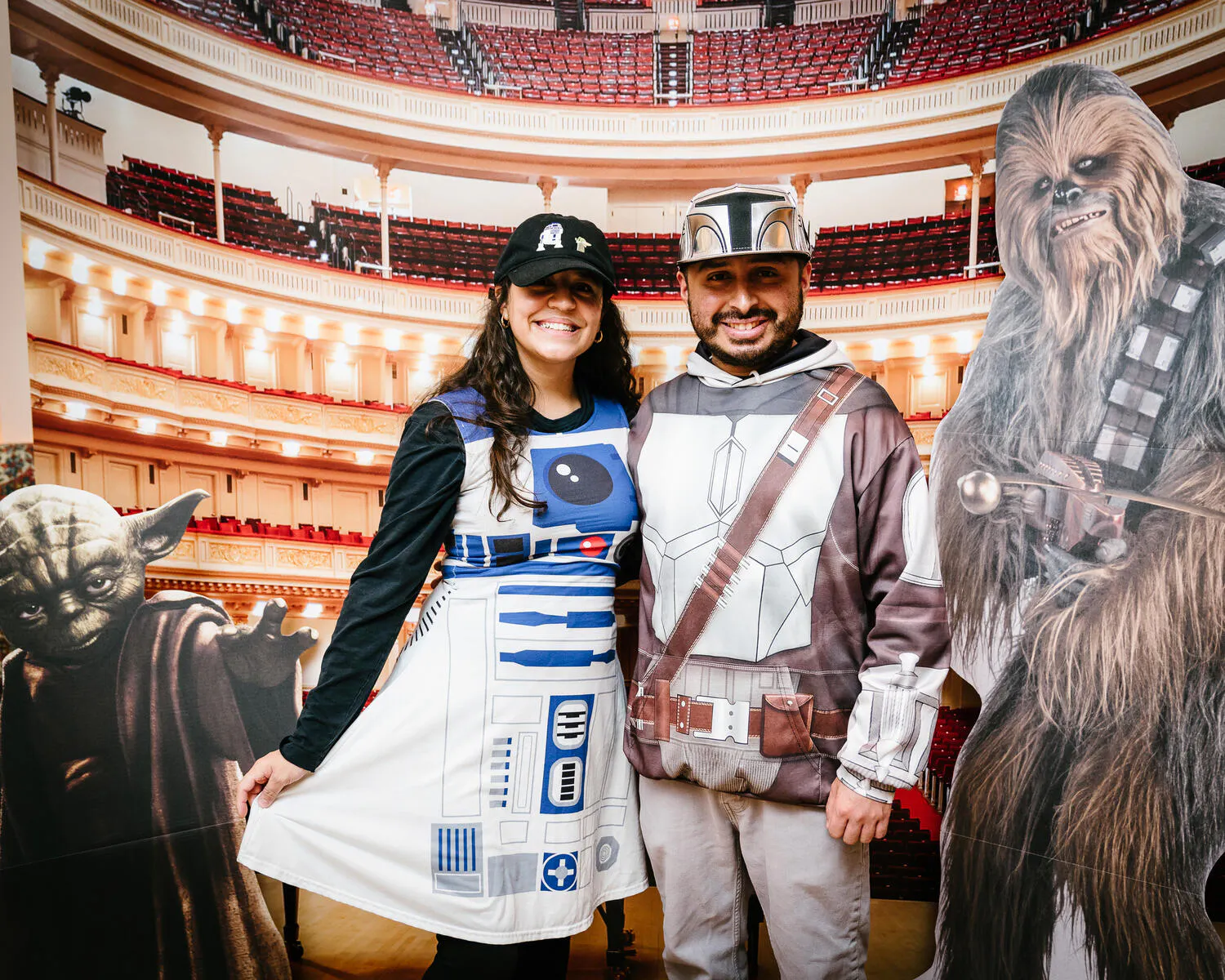 Two people pose in front of a Carnegie Hall backdrop with Star Wars characters beside them.