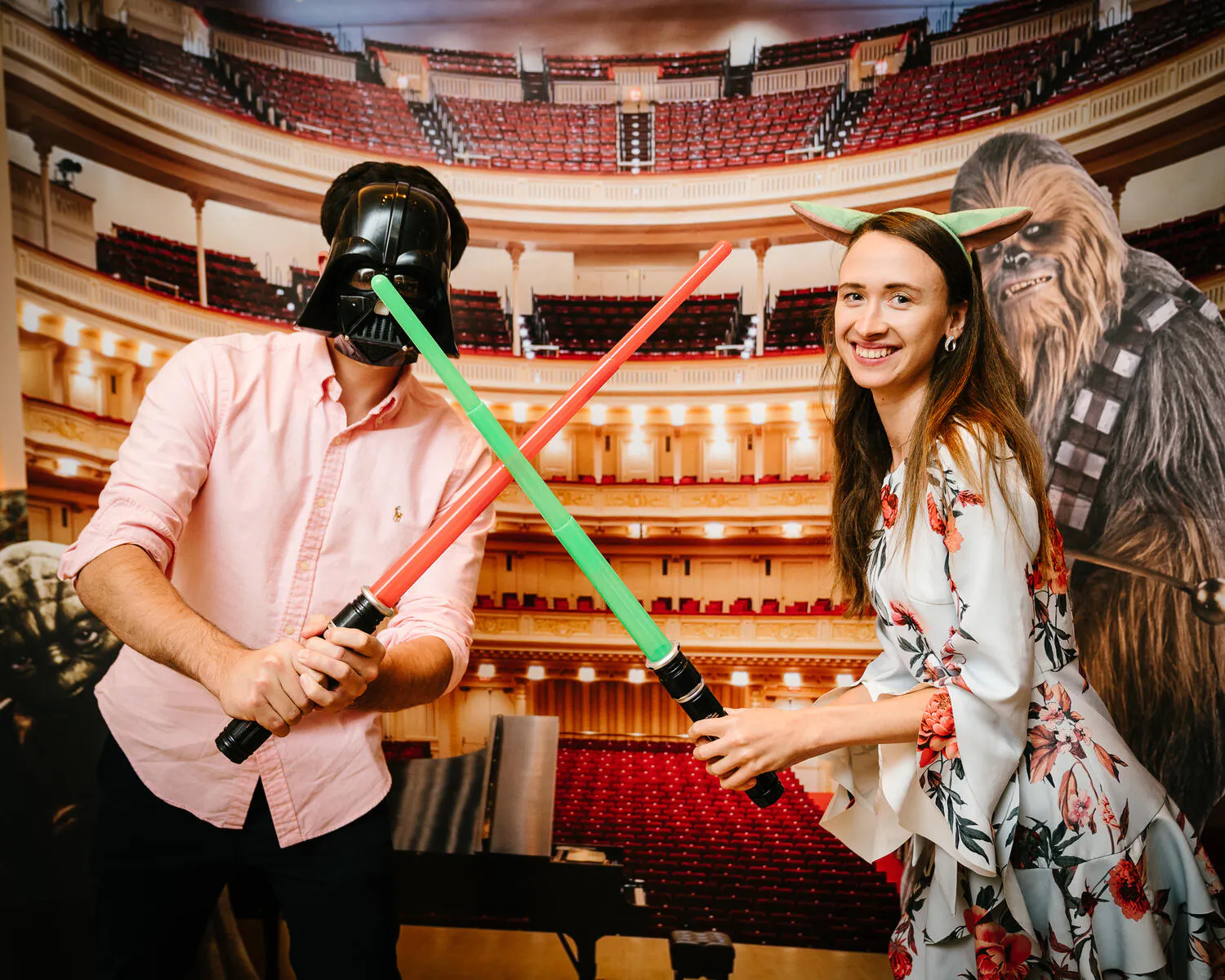 Two people hold green and red lightsabers in front of a backdrop of Carnegie Hall.