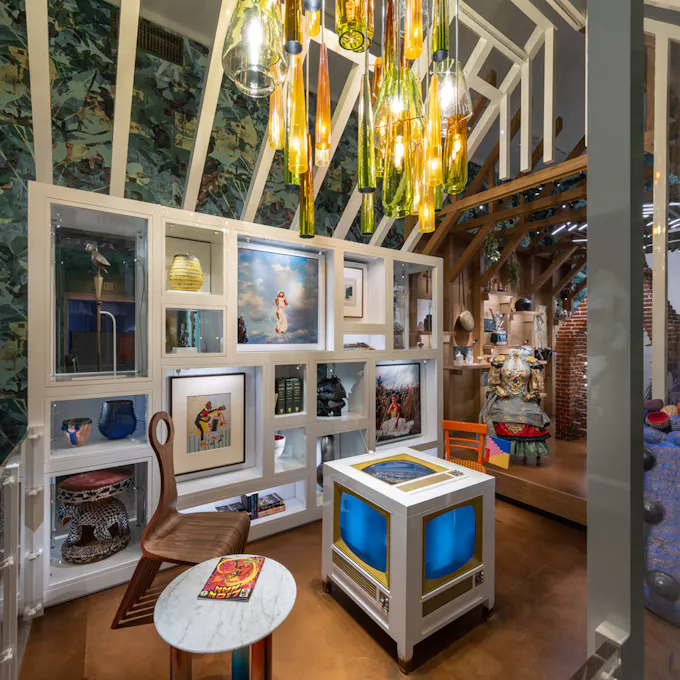 An art exhibition displaying a cabinet filled with paintings, vases, and books, and a table made to look like an old TV with a chandelier hanging overhead