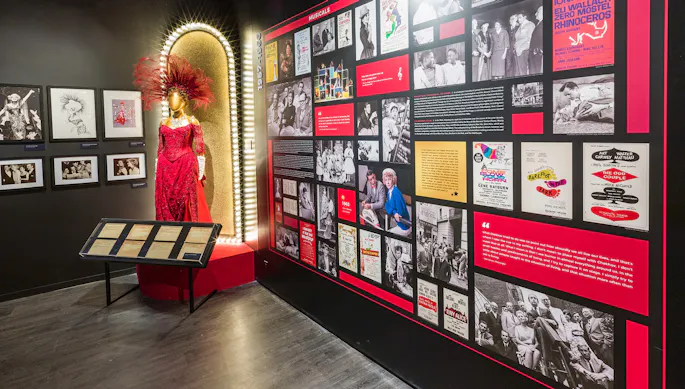 Exhibit of a mannequin wearing a red gown and feathered headdress next to a wall of photos and text