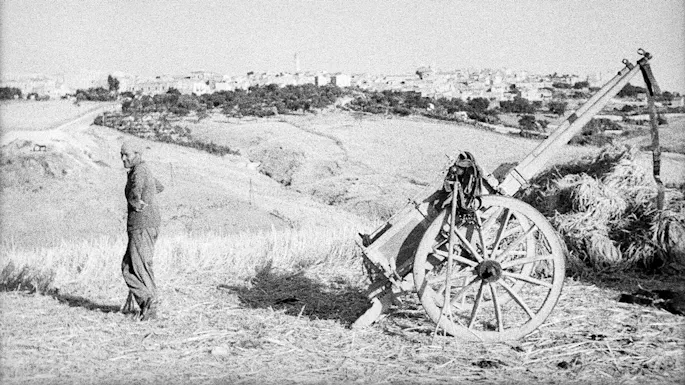 Grainy black & white photo of a man standing in a field across from a wagon, a city in the distance