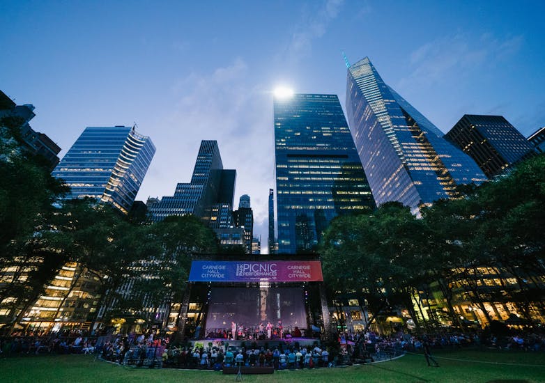 An audience gathers around a stage in Bryant Park at dusk.