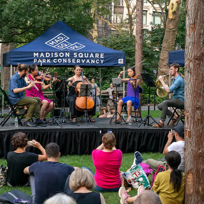 Ensemble Connect musicians perform on a raised platform in a park before a small crowd of people sitting on the grass