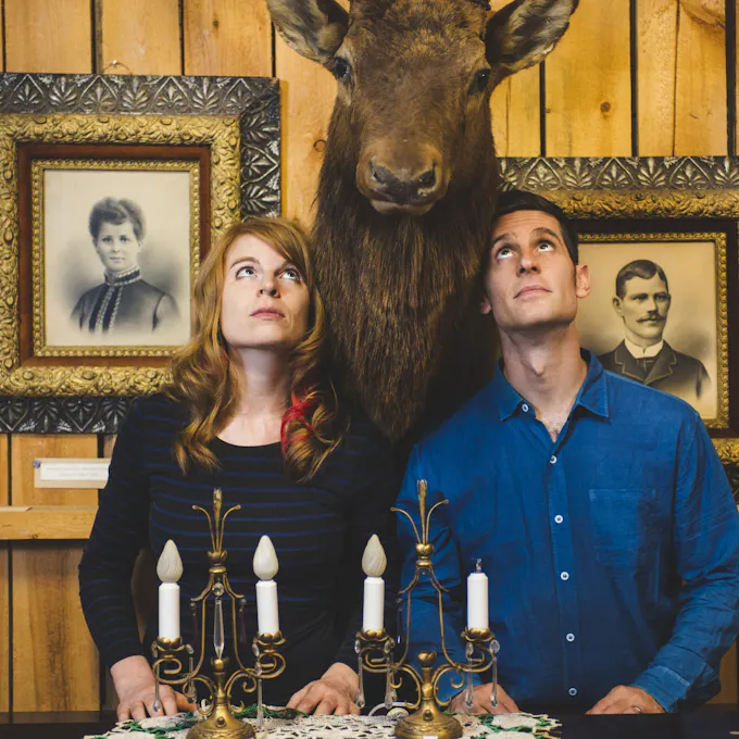 Jayme Stone and female bandmate standing on either side of a wall-mounted moose’s head
