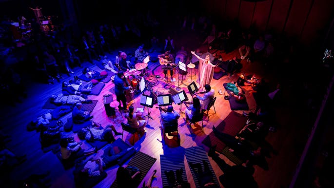 A woman stands with arms outstretched, circled by musicians and people lying on mats