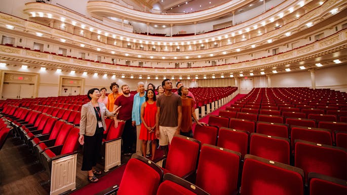 A group of people standing in the aisle of Stern Auditorium / Perelman Stage, with a tour guide pointing up to the balcony and speaking
