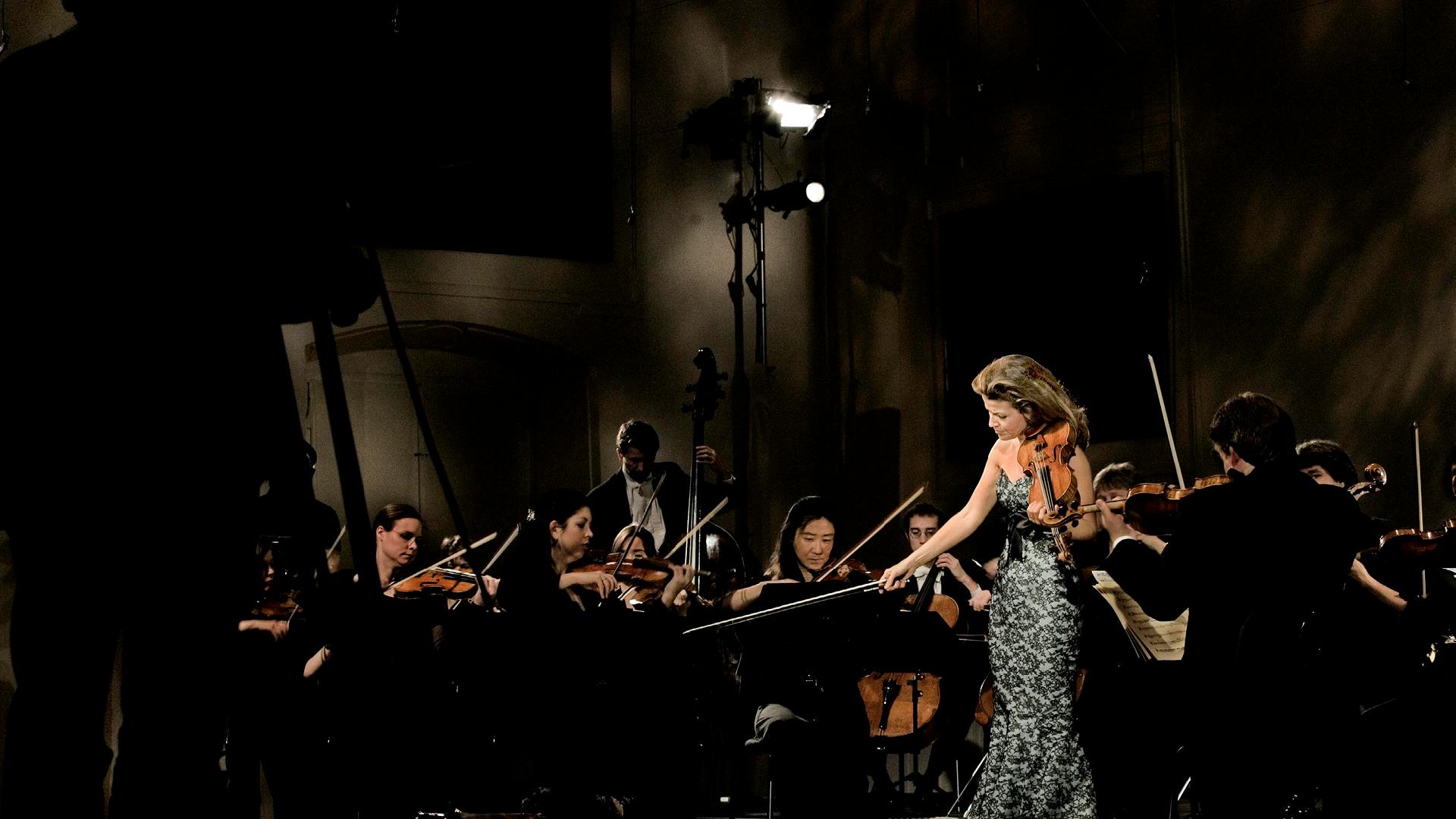 Anne-Sophie Mutter standing with a violin on stage in front of an orchestra.