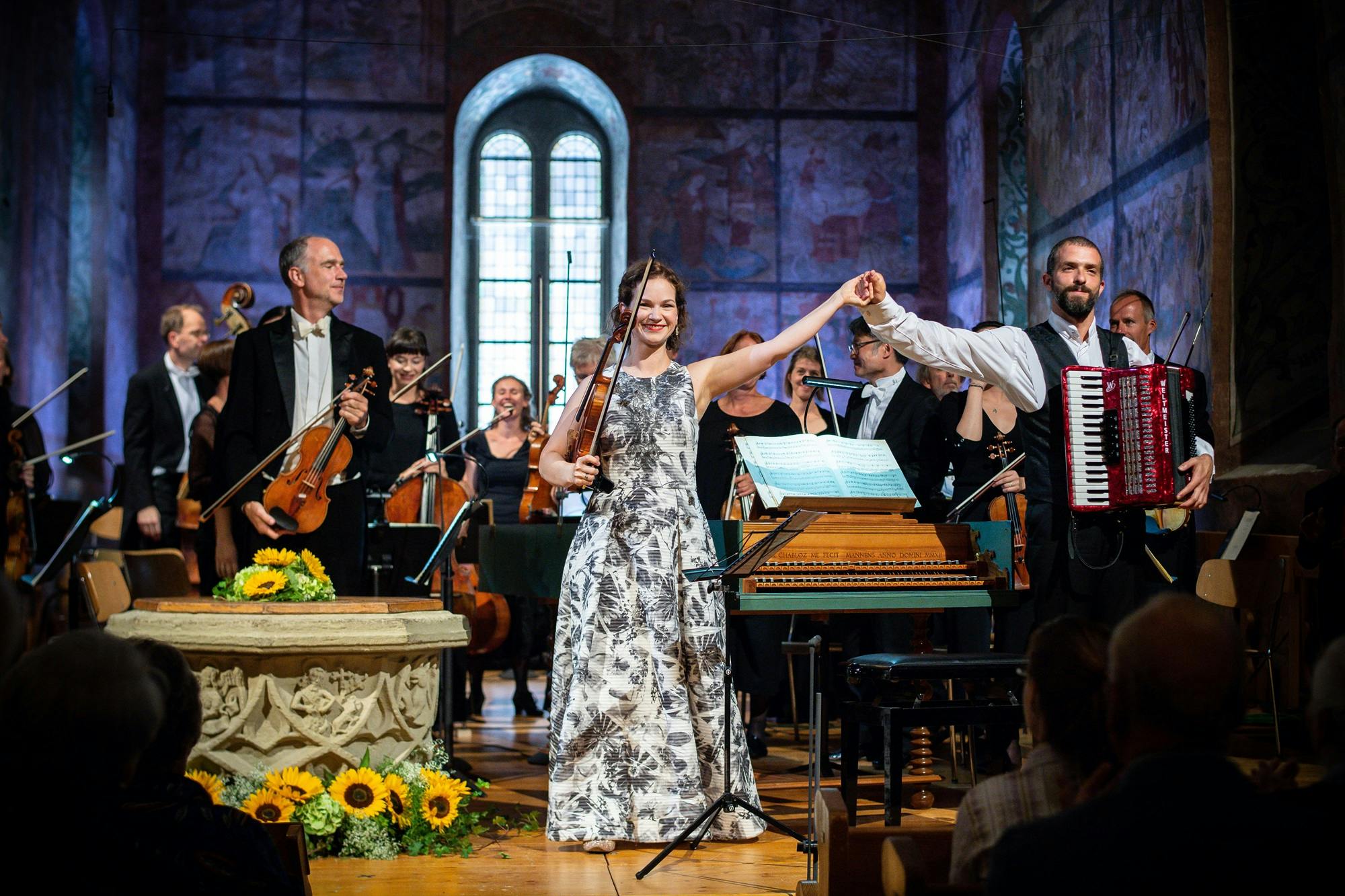Hilary Hahn and Omer Meir Wellber holding raised hands and standing with orchestra musicians for applause.