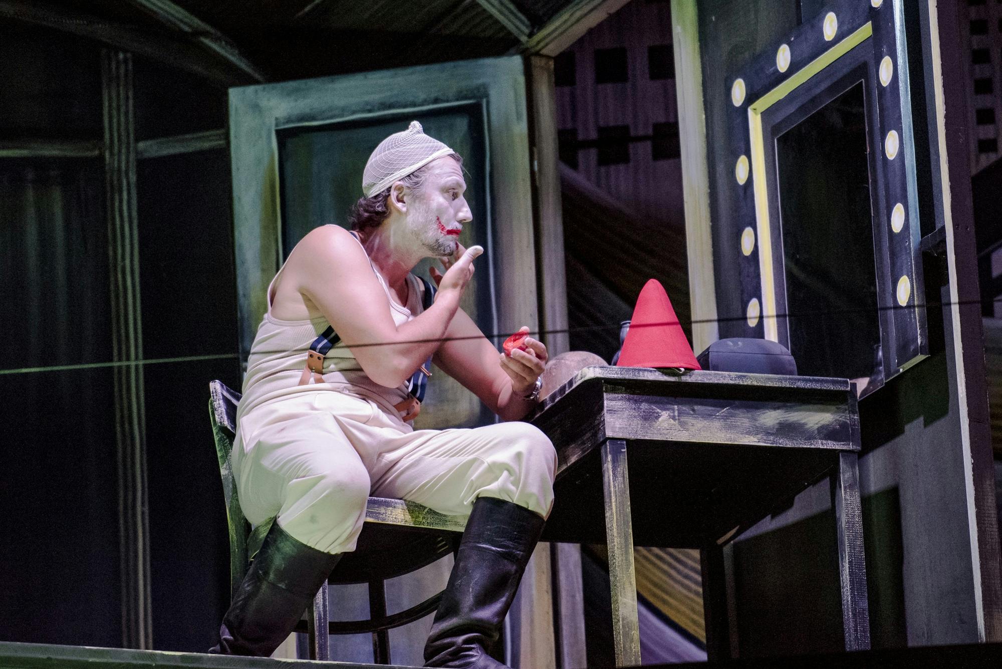 Jonas Kaufmann sitting in front of a mirror applying clown makeup.