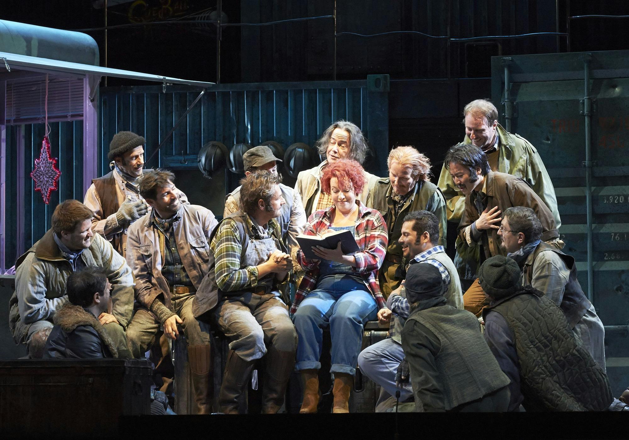 A group of performers gathered around Nina Stemme, who is reading a book.