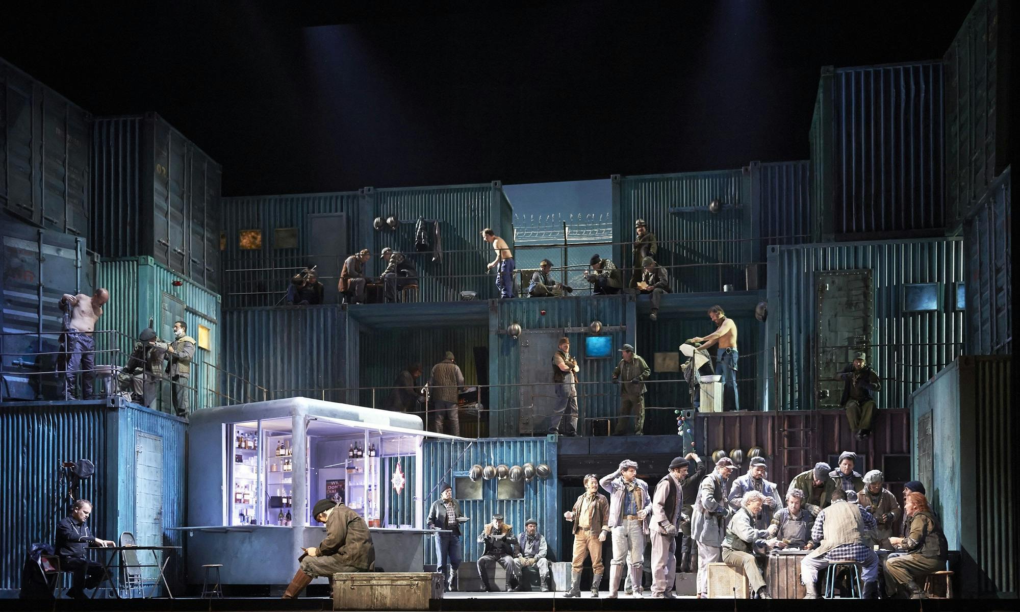 An ensemble cast on stage simultaneously depicting multiple scenes, including performers sitting at and gathering around tables, getting dressed, and eating food.