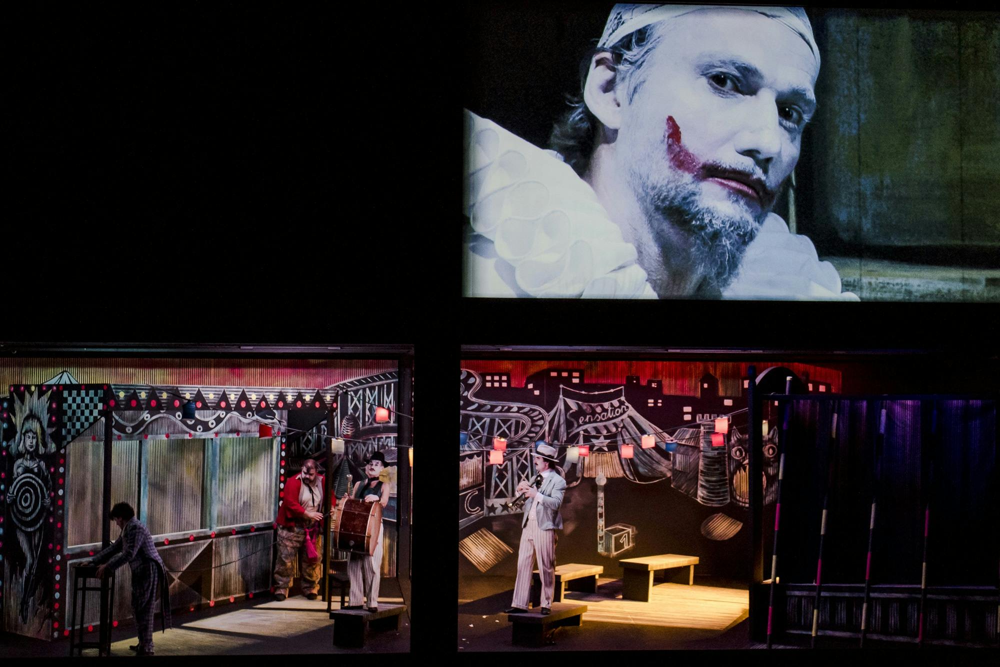 Performers dressed in colorful suits standing on stage, and a screen projection of Jonas Kaufmann in a clown costume displayed above.