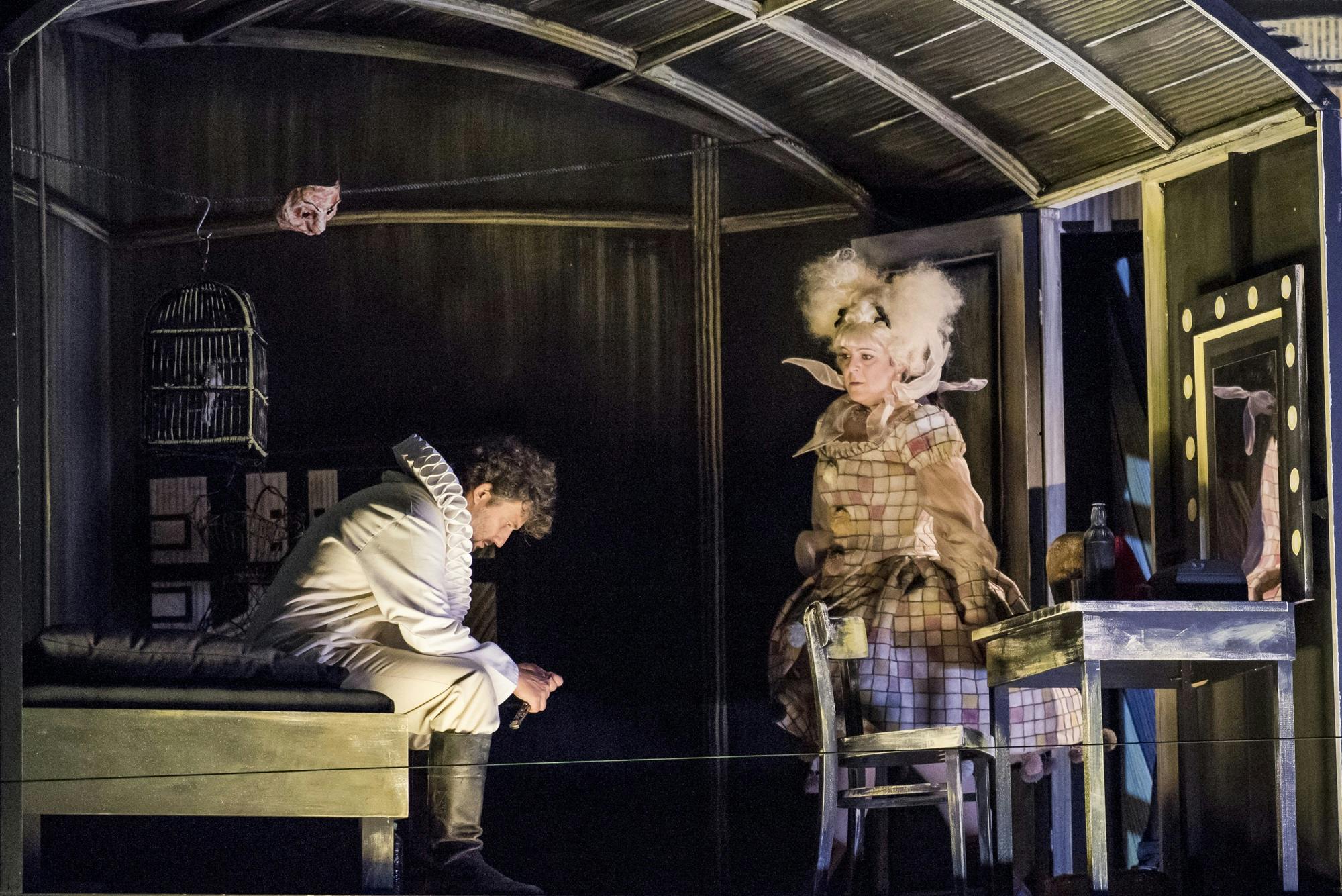 Jonas Kaufmann sitting on a bed, and Maria Agresta standing near the door.