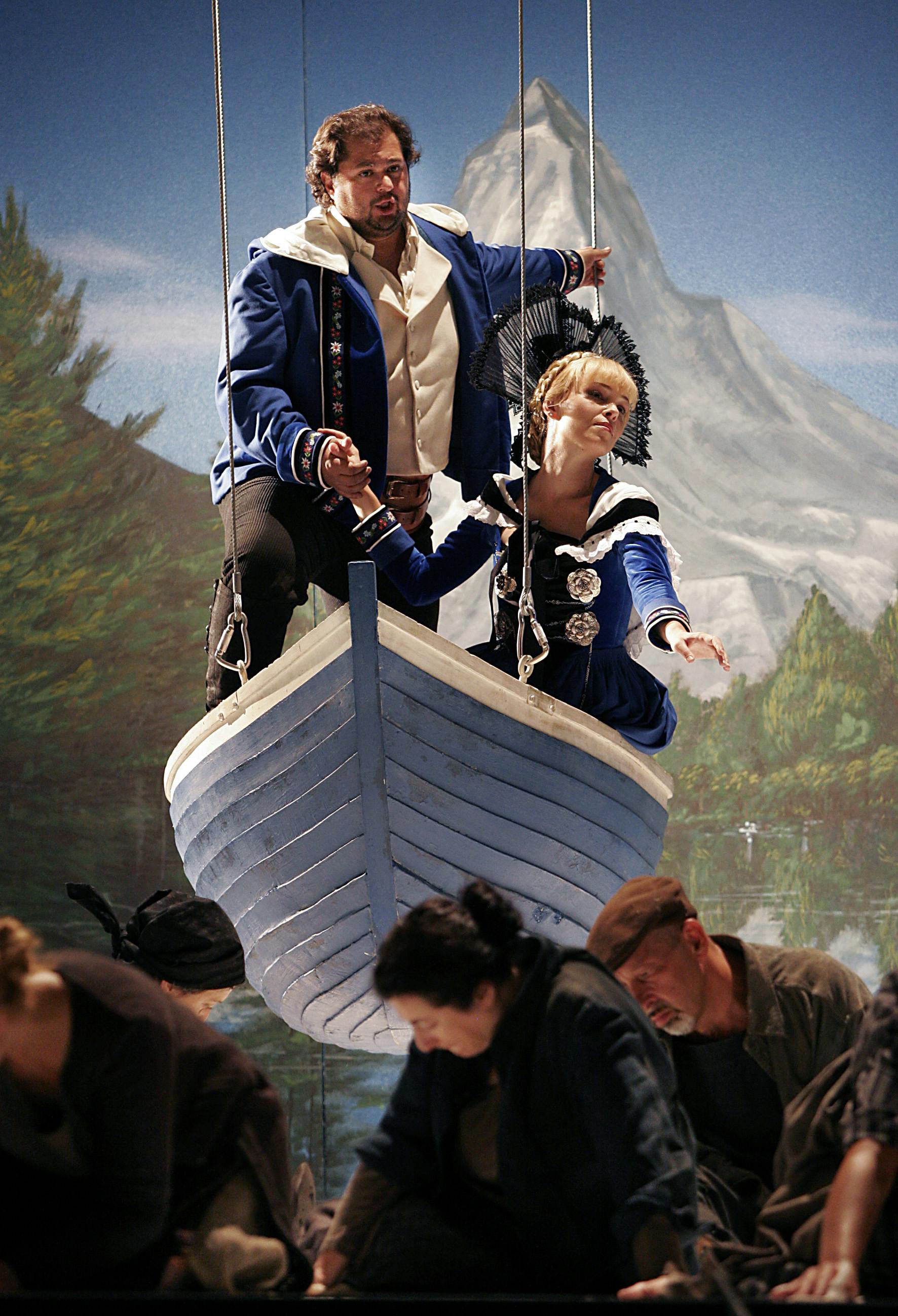 Two performers standing up in a suspended boat, with other performers kneeling on stage.