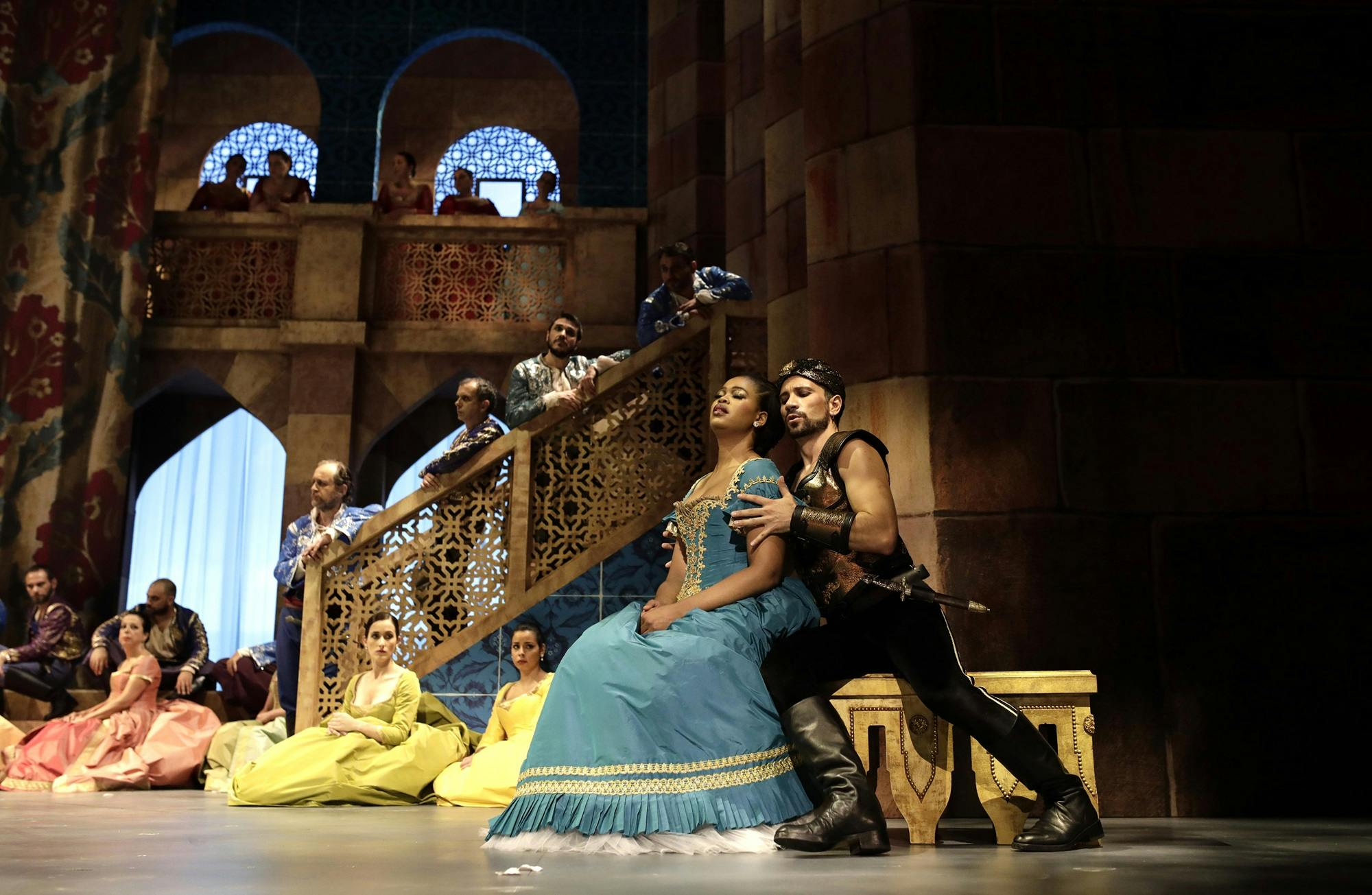 Pretty Yende sitting on a ledge, behind held by a peformer. Several performers sit behind on the ground, and others stand on the staircase and balcony in the background.