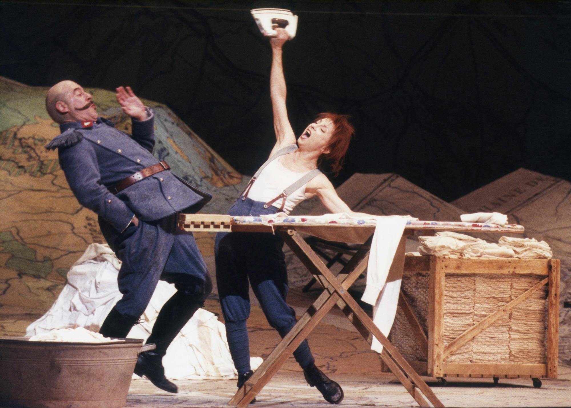 Natalie Dessay singing and ironing clothes, raising the iron in the air; a second performer leans back in surprise.